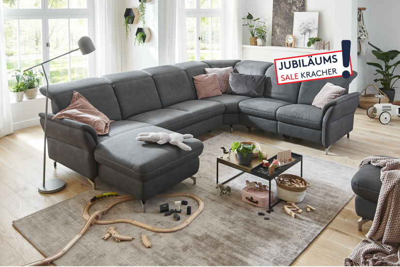 Polstergarnitur Senturo – Dunkles Ecksofa mit Hocker in realistischem Wohnzimmerambiente mit Teppich, Couchtisch und Deko.